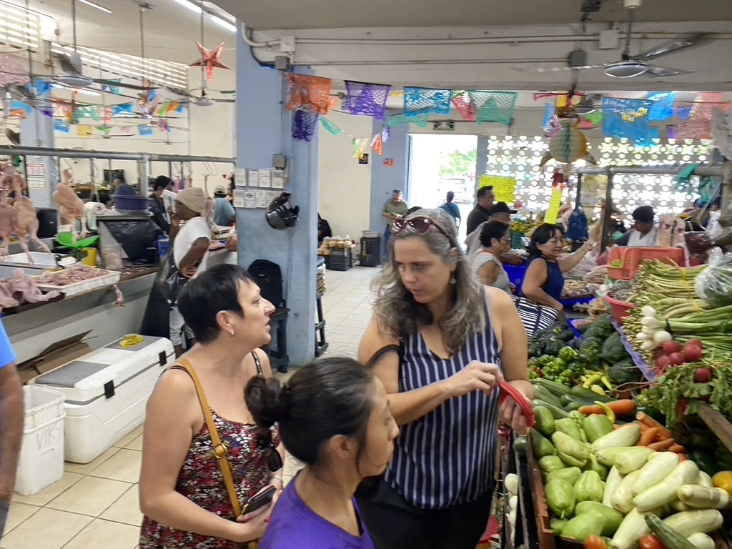 Françoise y Yannick conversando en español con una vendedora mientras seleccionan ingredientes frescos en un mercado local de Mérida.